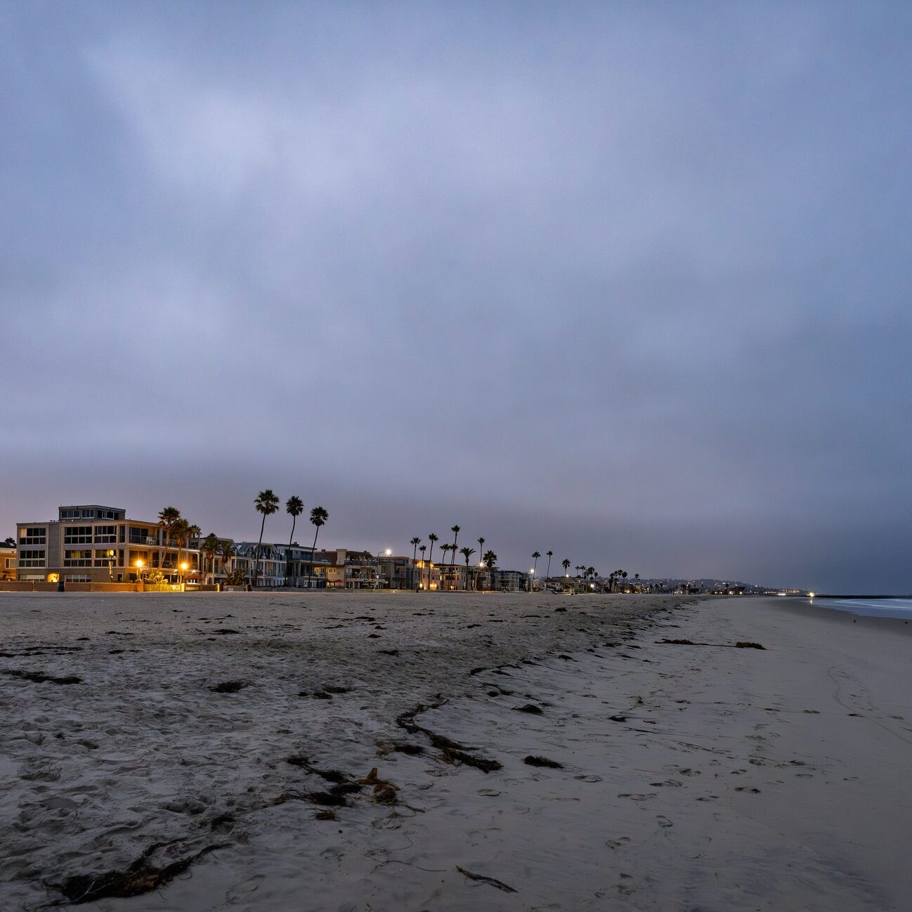 A wide, serene photograph of the beachfront in Mission Beach, California, captured at twilight. The low, moody clouds and deep blue sky hang over the scene as the last light of the day fades. The lights from the beachfront homes and streetlights begin to glow, creating a gentle illumination that contrasts with the shadowy palm trees. This long-exposure shot captures the peaceful, transitional moment between day and night on the coast. High quality photo